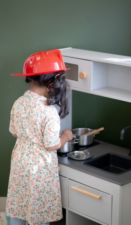 Une enfant, de dos, cuisine dans la cuisinette avec un casque de pompier